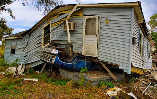 Photograph of a wrecked house on top of an upside down blue pickup truck.