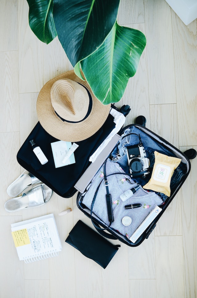 Suitcase with sun hat and clothing laid on the floor with the large leaves of a tropical plant hanging overhead. Inside and around the luggage are vacation items - A 35MM camera, sunblock, sandals, body wipes, and notepad.