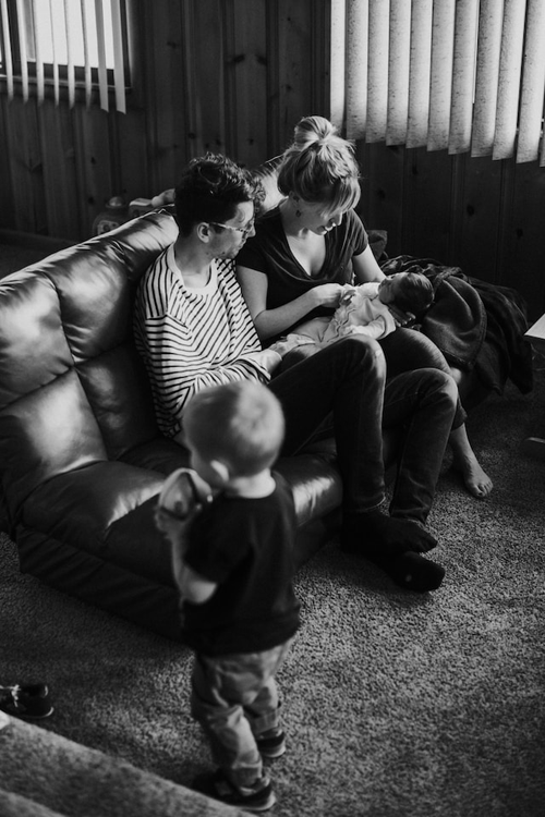 A black and white image of a Caucasian family of 4. The father and mother are sitting on a leather loveseat holding an infant while a toddler walks past them holding a toy.