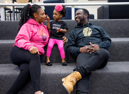 Image of an African-American mother, child, and father. The child is between the parents and all are smiling. The child is wearing a bright pink shirt with mini boss written on it. The child is also pointing at the mother.