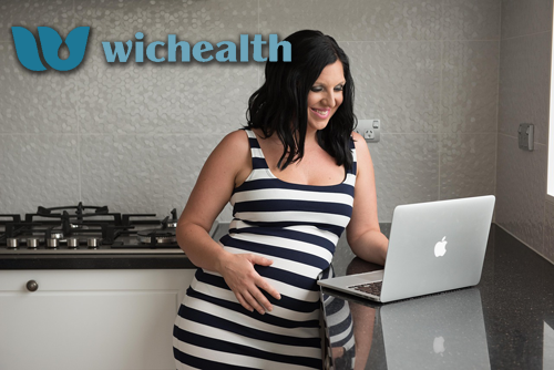 Pregnant woman browsing on a MacBook while standing in the kitchen. There is some type and a logo that reads wichealth
