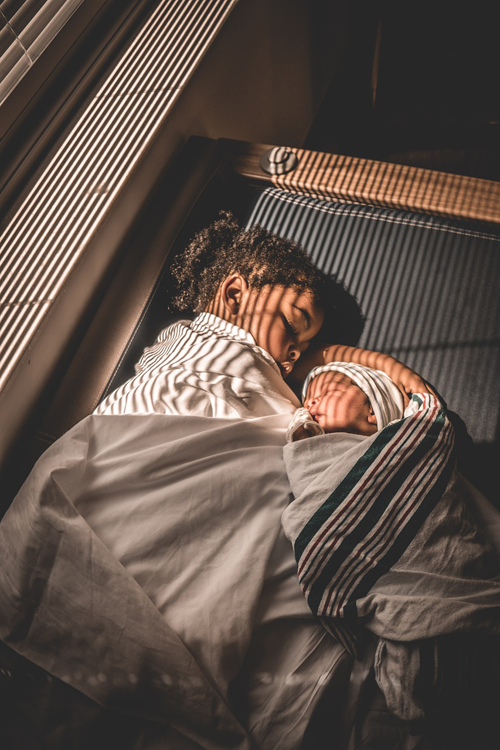 Image of 2 sleeping boys. One is a toddler, the other an infant. They are wrapped in blankets with sun peeking through the blinds of the room.