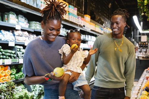 Imagen de una madre, un hijo y un padre afroamericanos en la sección de productos agrícolas de una tienda de comestibles. La madre sostiene al niño mientras inspecciona una pieza de fruta.